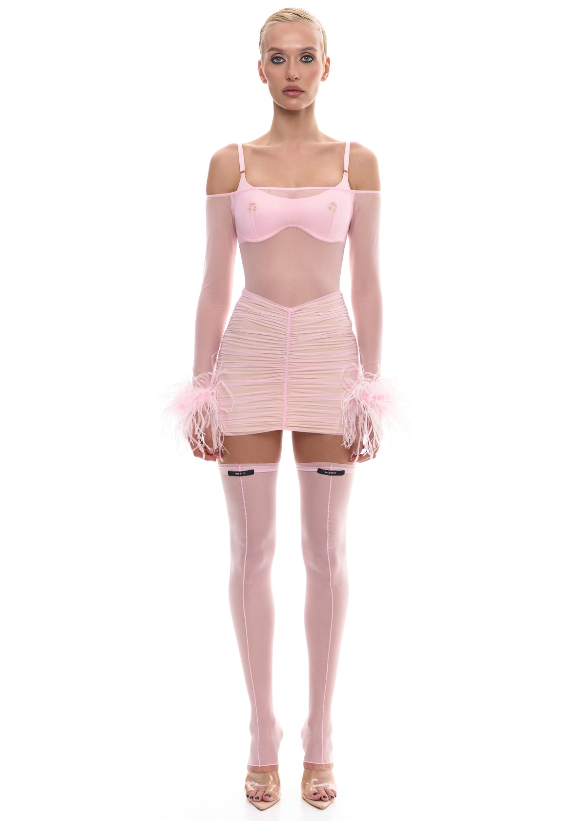 На моделі рожевий комплект із атласу з імітацією пірсингу (колір фурнітури золото), прозорий рожевий боді з страусинним пірʼям, рожева спідниця із латвійської сітки з м'яким еластичним підкладом та рожеві панчохи еластичні з латвійської сітки без носочку  зі швом спереду та ззаду із брендованою біркою. 